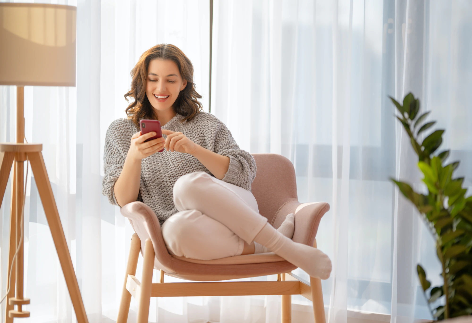 Happy casual beautiful woman is using a phone sitting on armchair at home
